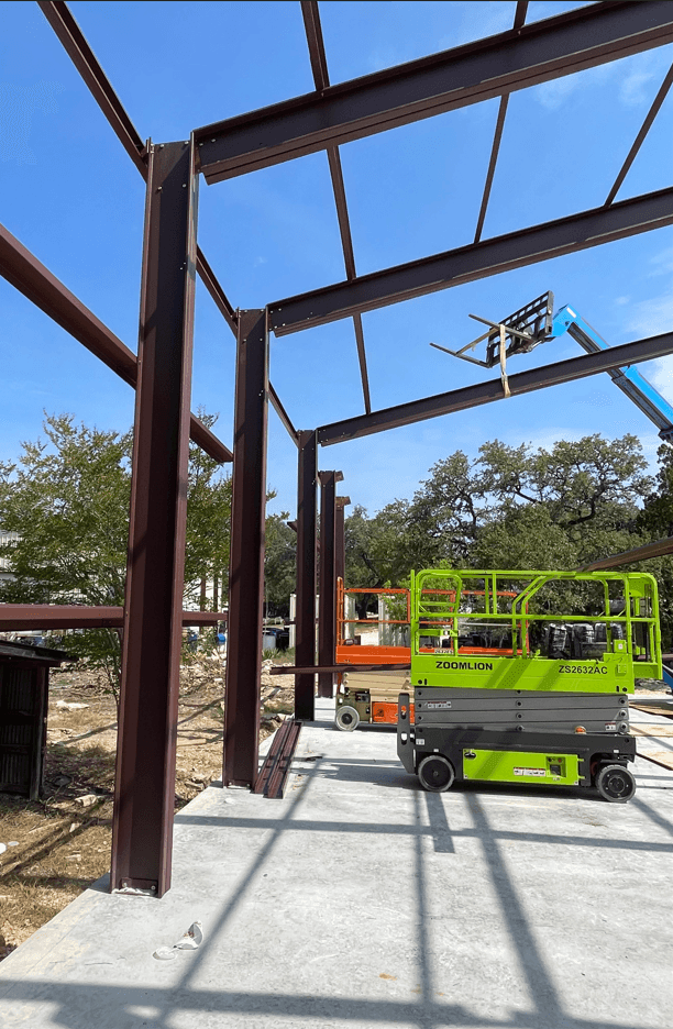 Construction site with steel beams and a green lift on concrete foundation under clear blue sky.