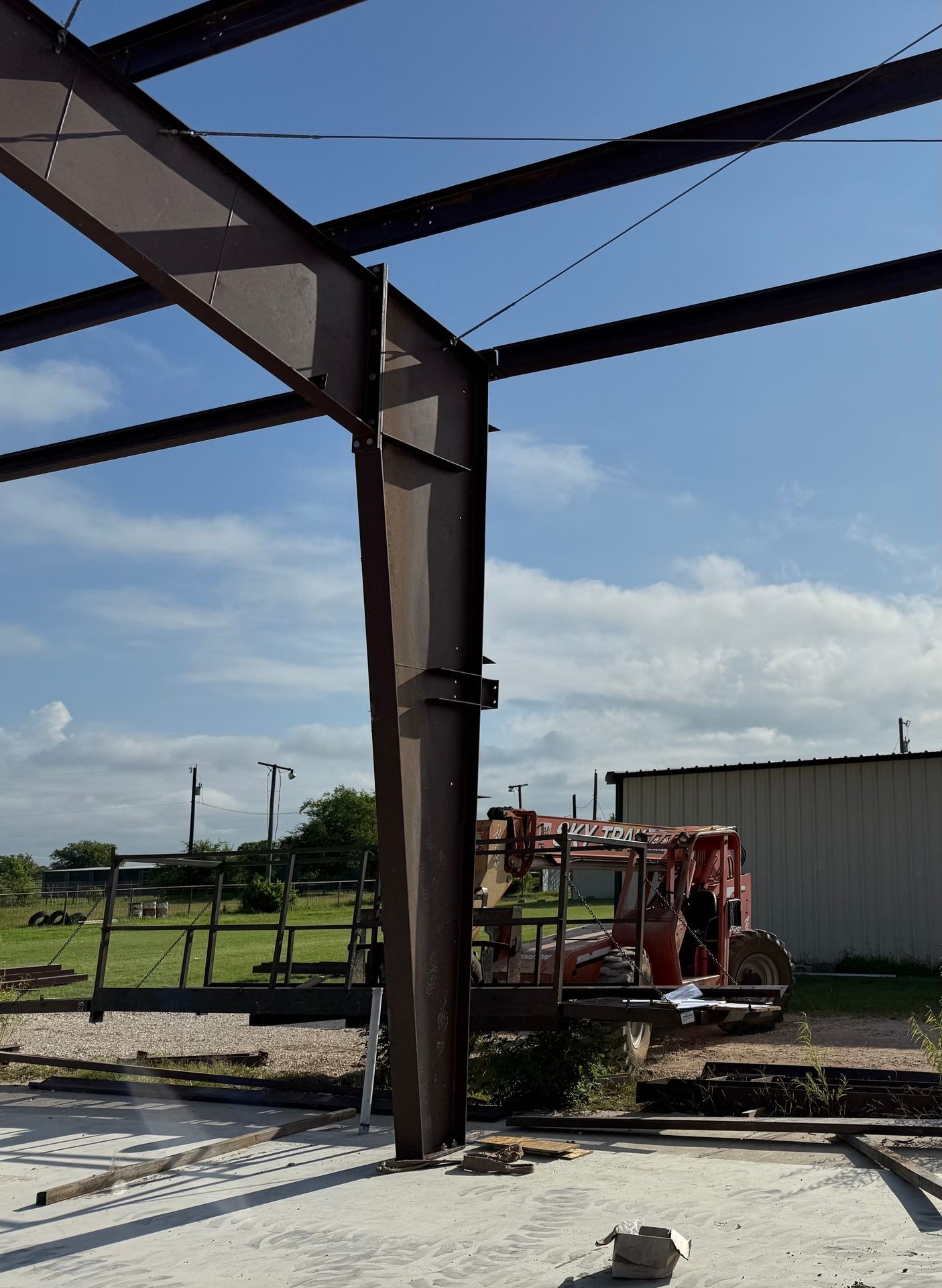 Construction site with metal beams, forklift, and storage building under a blue sky with clouds.